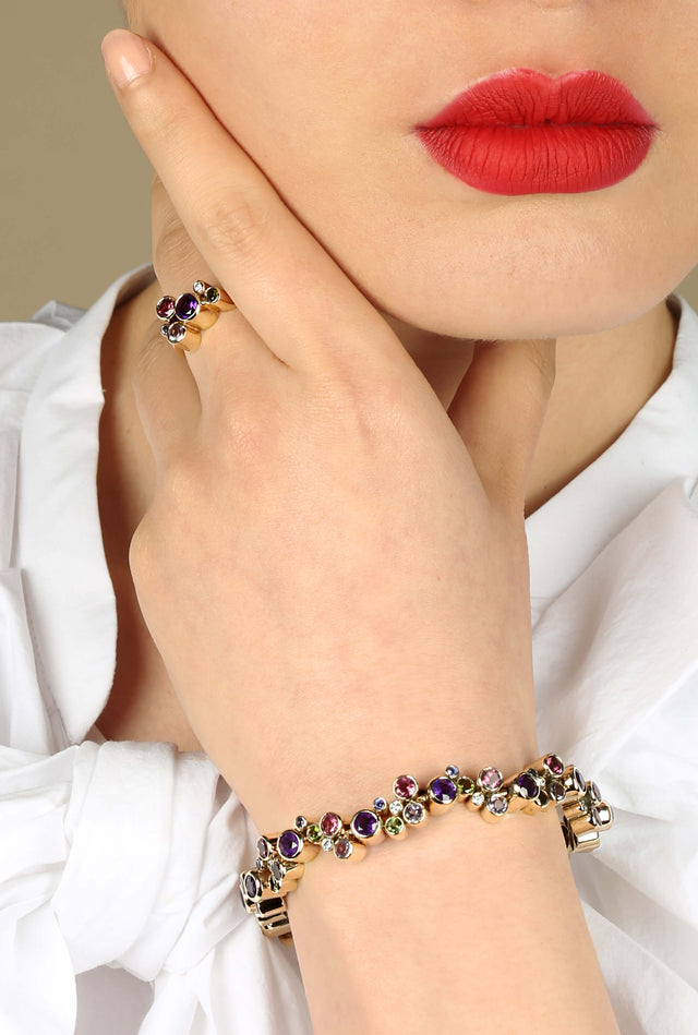 A women is holding her neck. She has bright red lips and a white top. She is wearing a Roundhaus bracelet and ring made in 18 carat yellow gold and brightly coloured precious gemstones.